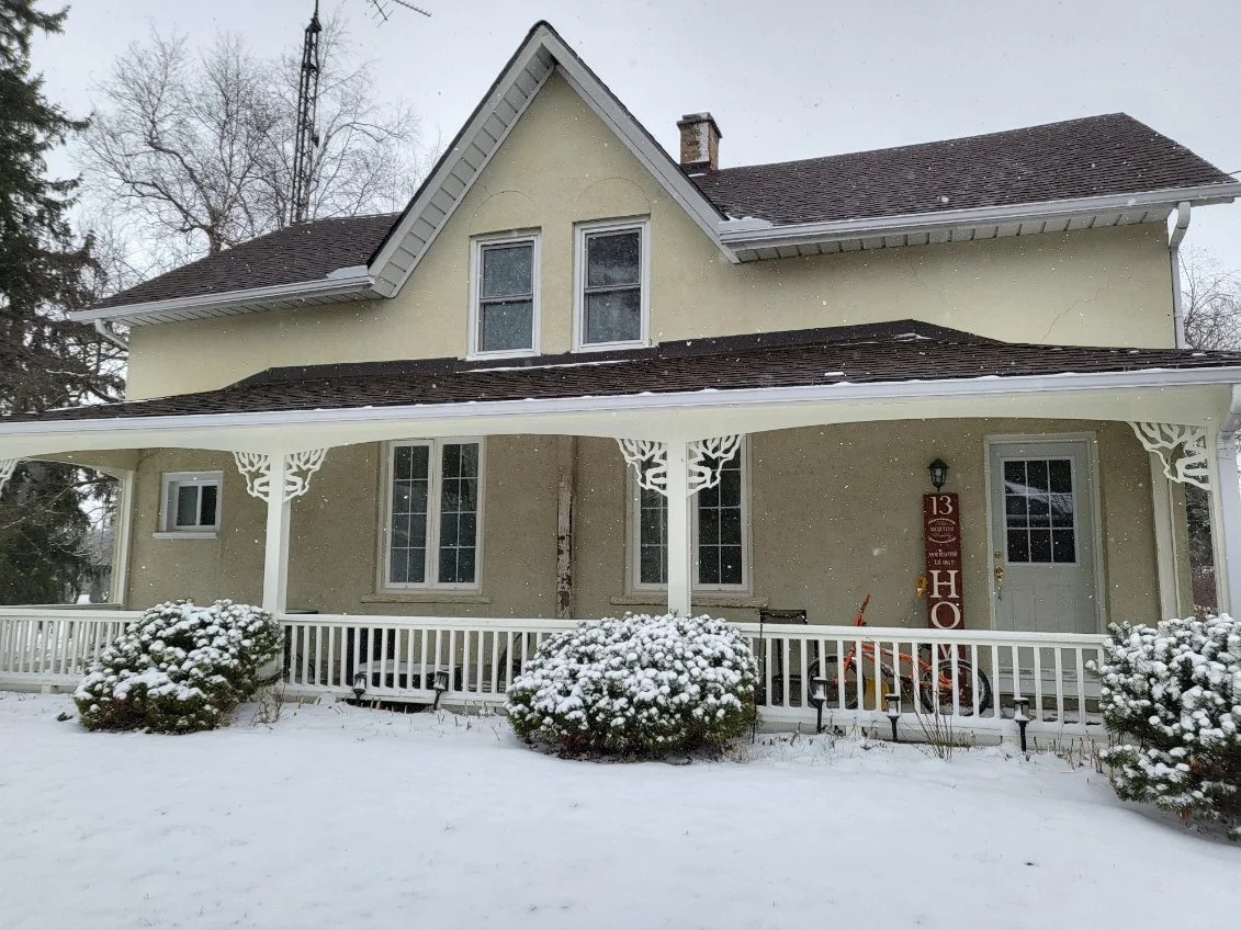 A two-story house with a covered front porch, light yellow exterior, and snow-covered yard and bushes in winter.