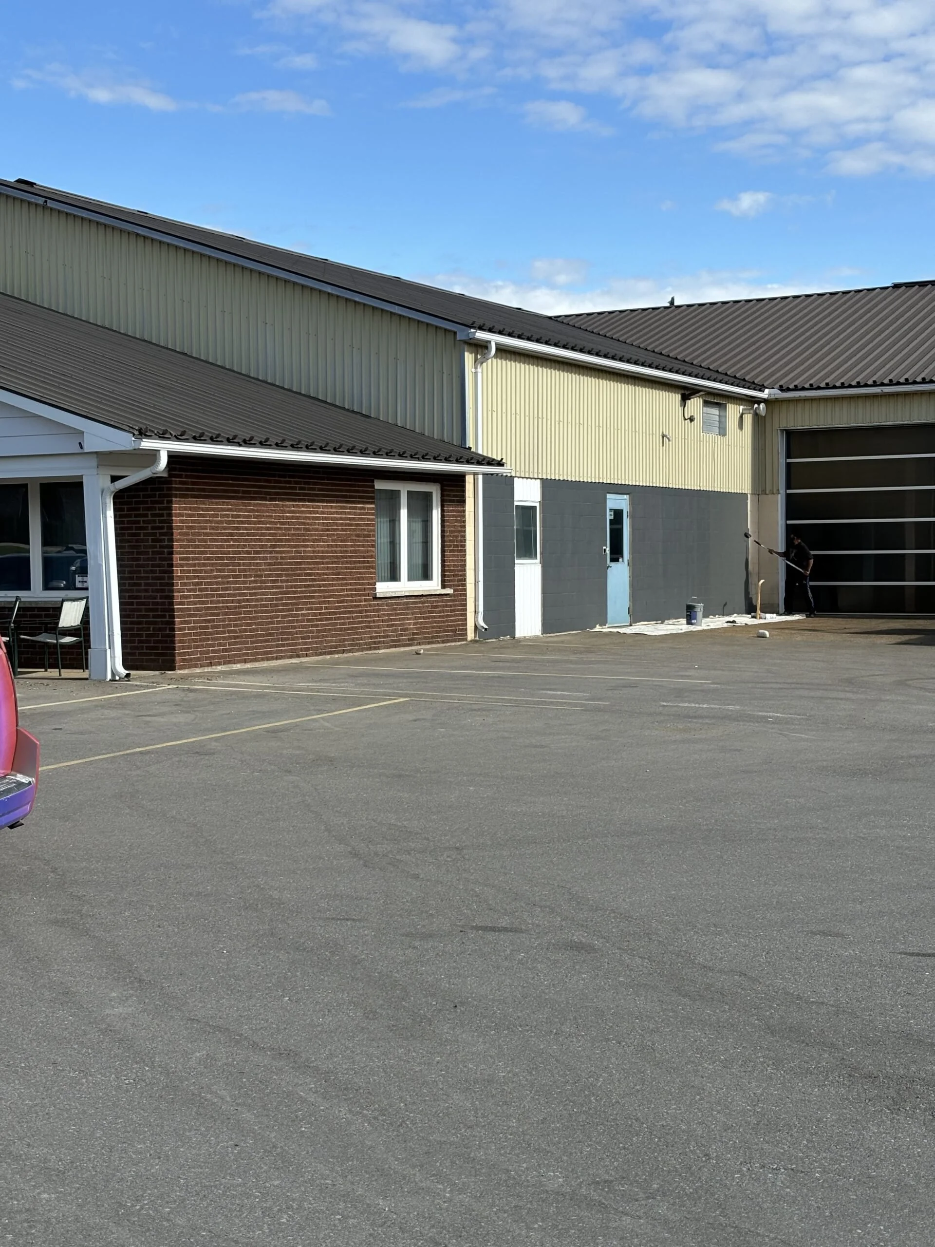 A building with a brick wall, windows, and a garage door, with a parking lot and a person painting the wall.