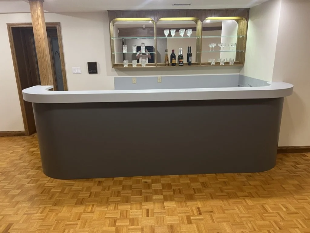 Empty home bar with a curved gray counter, wooden shelves with glassware and bottles, and a hardwood floor.