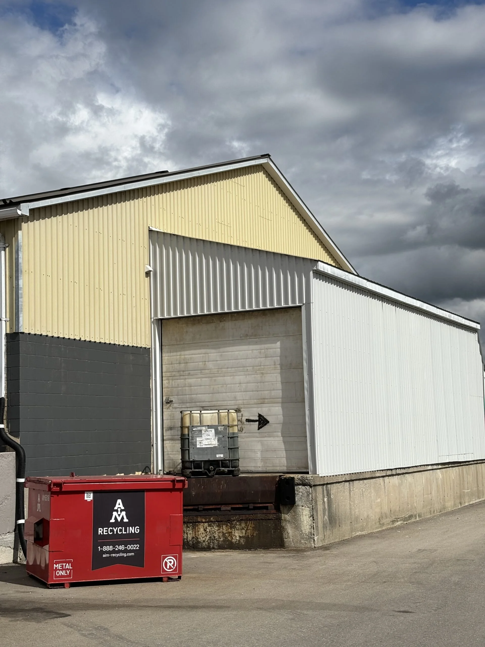Industrial warehouse building with a yellow and white metal exterior, a large garage door, and a red recycling bin labeled 'AM Recycling,' located on a concrete platform under a cloudy sky.