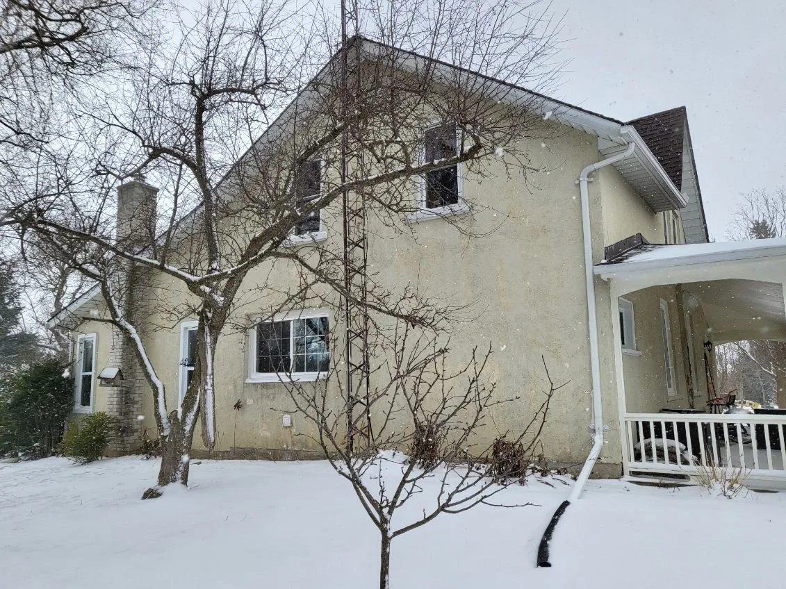 A two-story house with a light yellow exterior, snow on the ground, and leafless trees in front. The house has several windows, a chimney, and a covered porch area on the side.