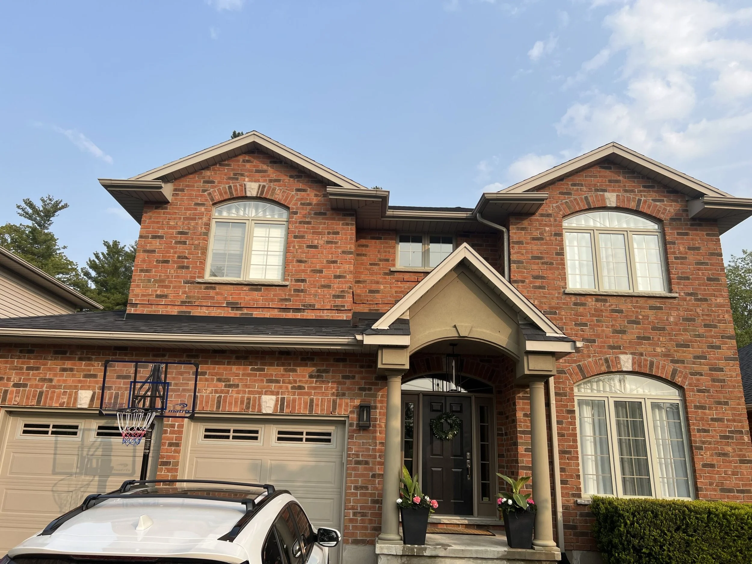 A two-story brick house with a front porch, decorated with plants in pots, with large white-framed windows, a black front door with a wreath, a driveway with a white car, and a basketball hoop nearby, under a blue sky with some clouds.