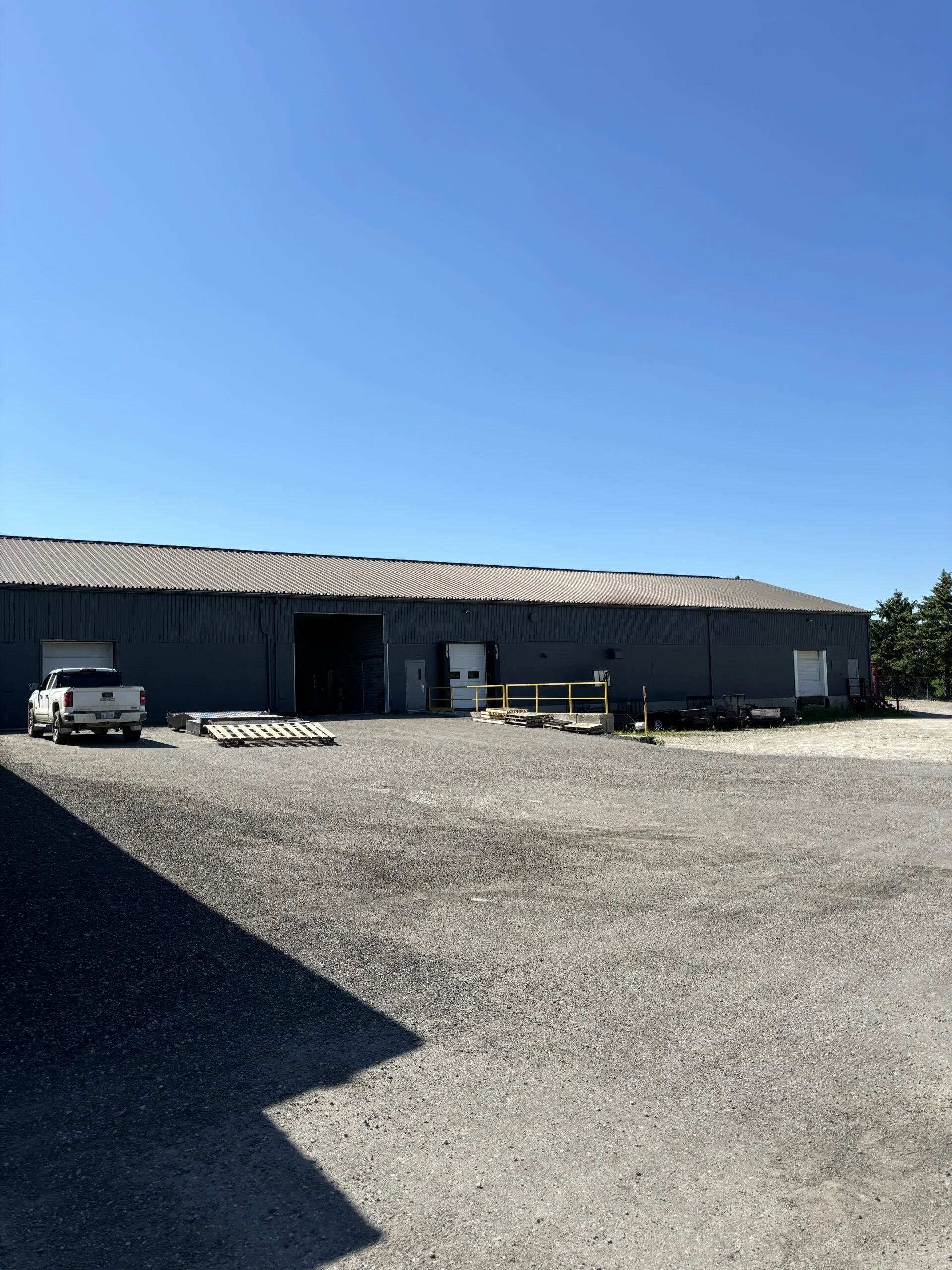 A large gray industrial warehouse with a metal roof, a loading dock, and a parking area with a white pickup truck. Clear blue sky and some greenery in the background.