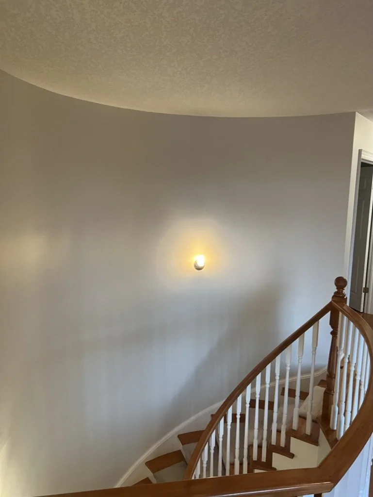 Interior view of a staircase with a curved wooden handrail and white balusters, a wall with a single wall-mounted light fixture, and a doorframe visible on the right side.