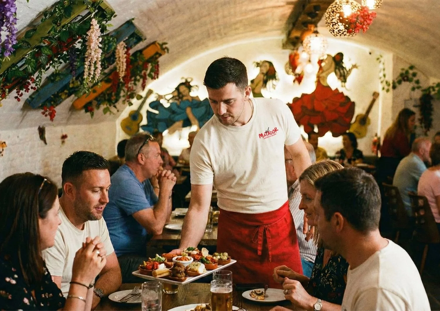 A waiter placing a tray of assorted tapas on a table in a lively Spanish restaurant with colorful decorations and murals of dancers in traditional costumes.
