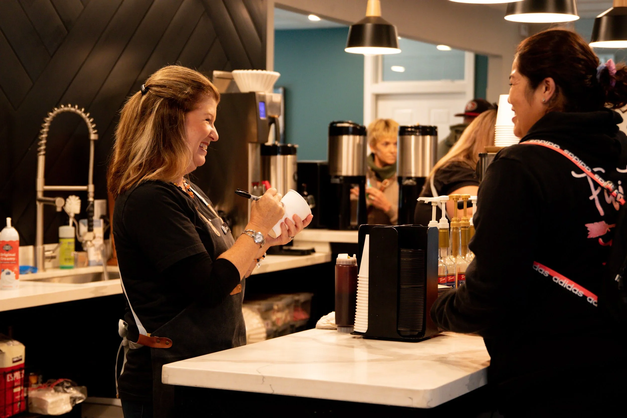 Two women consulting at a cafe counter, one preparing a takeaway coffee with a marker, smiling and engaging with the other, in a modern café setting.