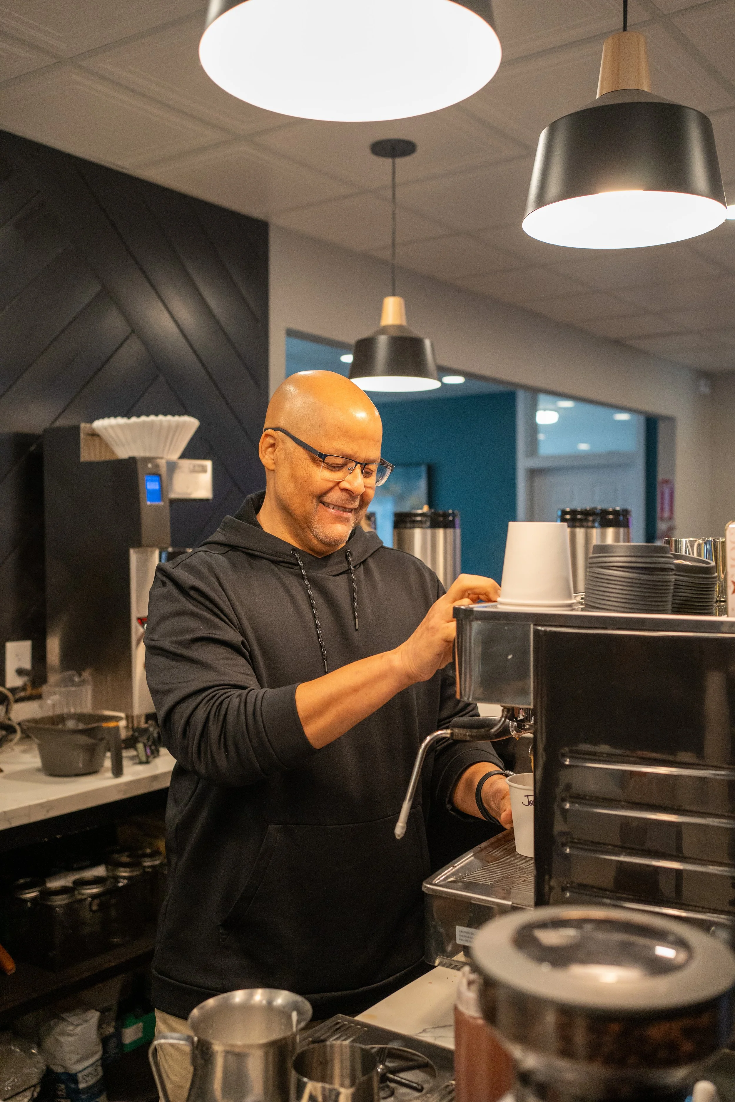 A man with glasses and a black hoodie making coffee behind a coffee machine in a modern cafe.