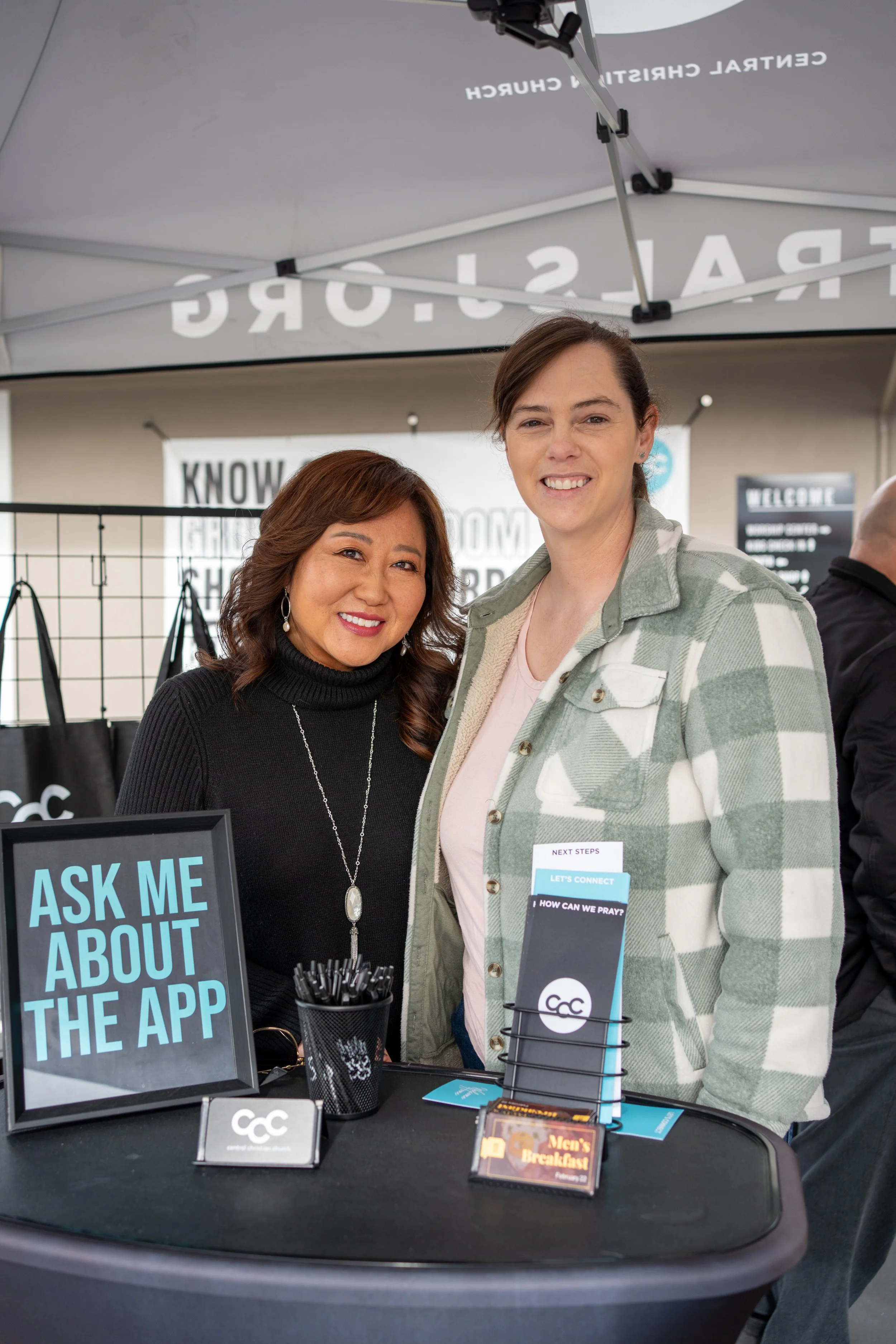 Two women standing behind a table at a booth, smiling at the camera. The table has a sign that says, 'Ask me about the app,' along with pens and brochures.