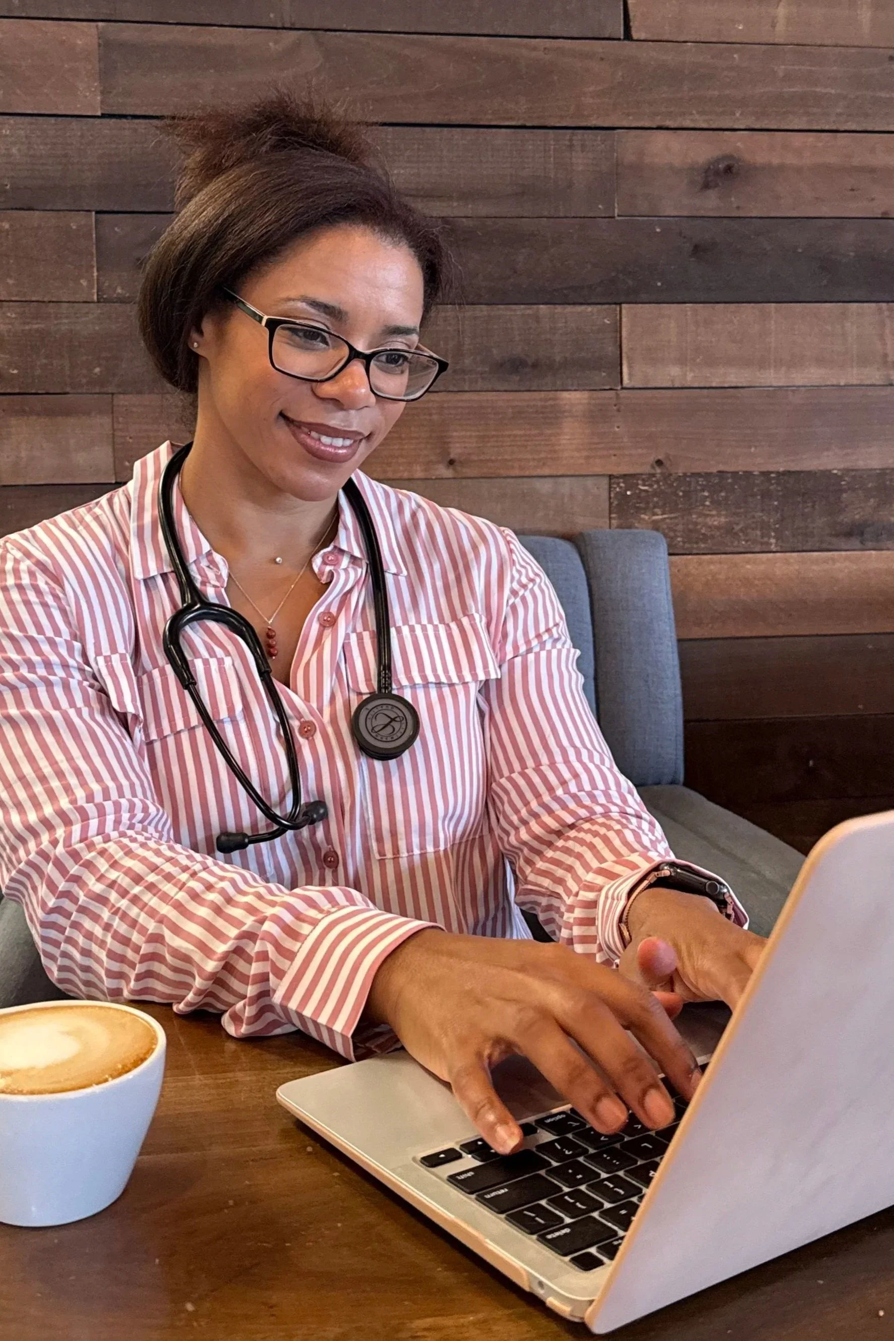 A smiling woman with glasses, wearing a striped pink and white shirt, with a stethoscope around her neck, working on a laptop at a cafe table, with a cup of coffee nearby.