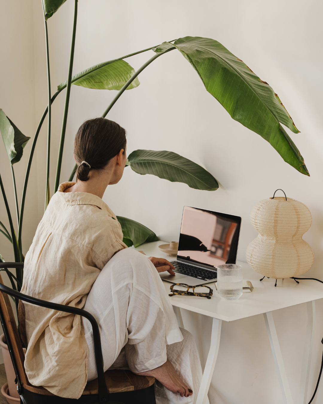 A woman with brown hair tied back, sitting on a chair at a white desk, working on a laptop. Large green plant leaves are behind her, a glass of water, a pair of glasses, and a decorative paper lantern are on the desk.