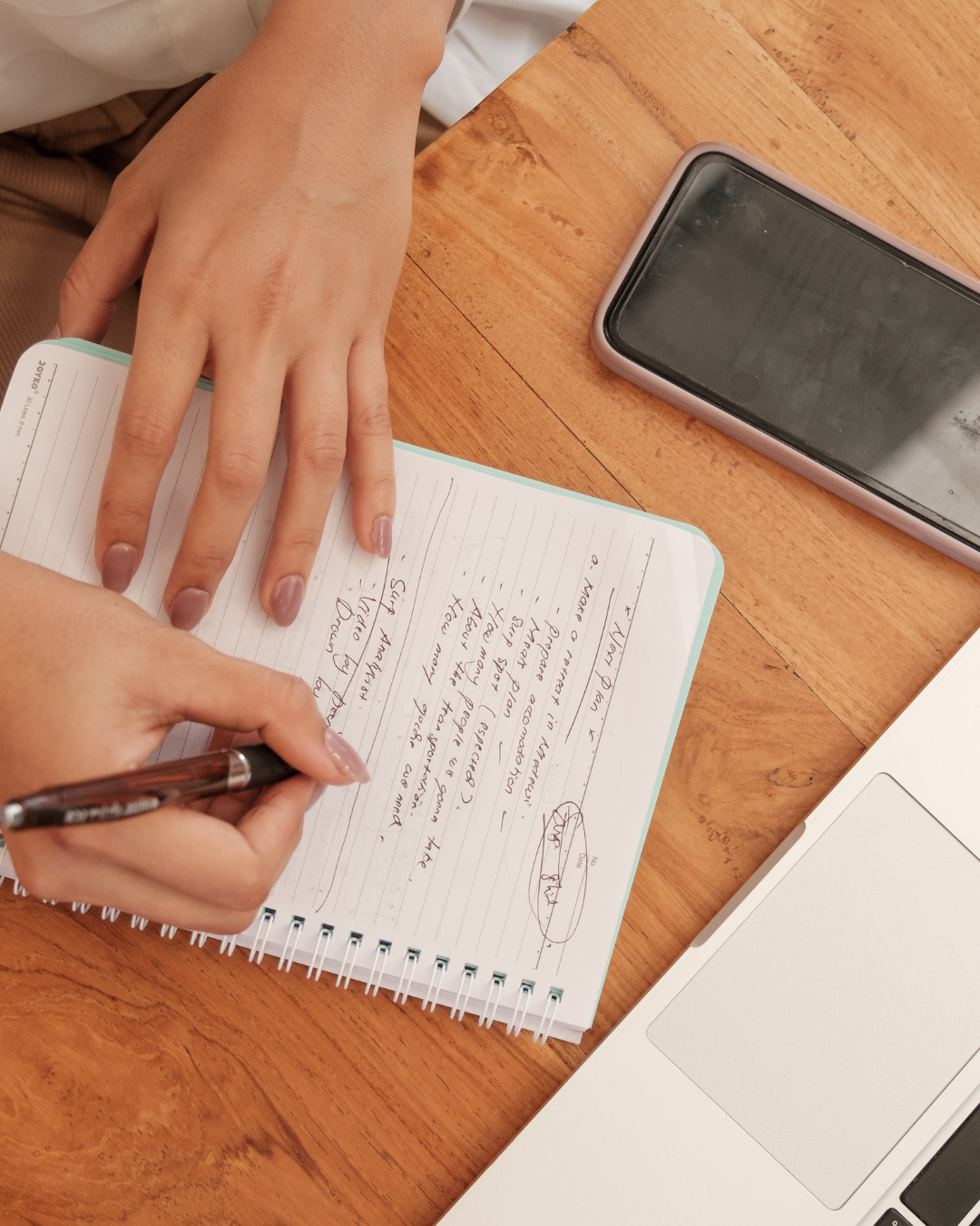 A person writing in a notebook with a pen, on a wooden table, with a smartphone and a laptop nearby.