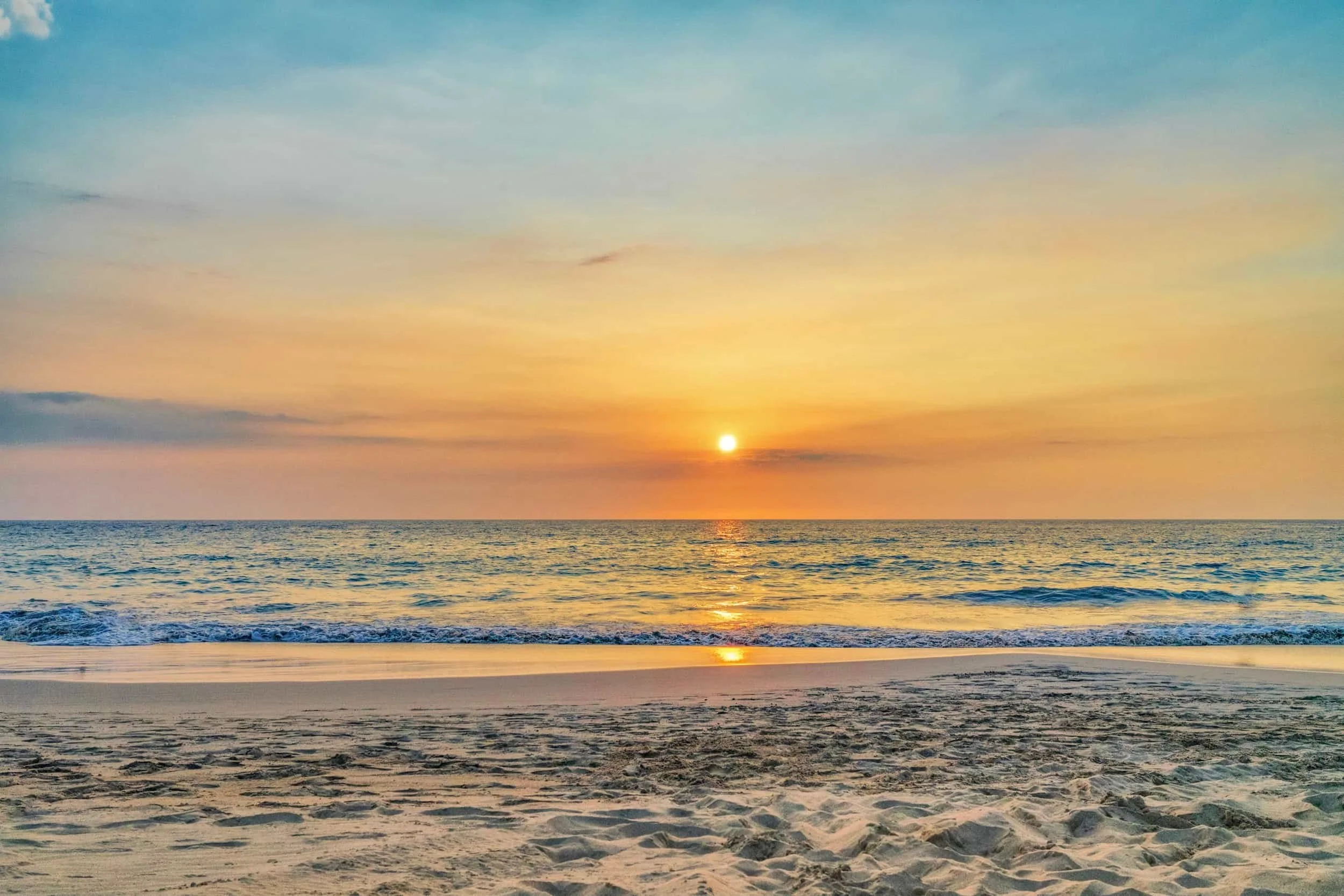 Sunset over the ocean with a sandy beach in the foreground and a colorful sky with light clouds.