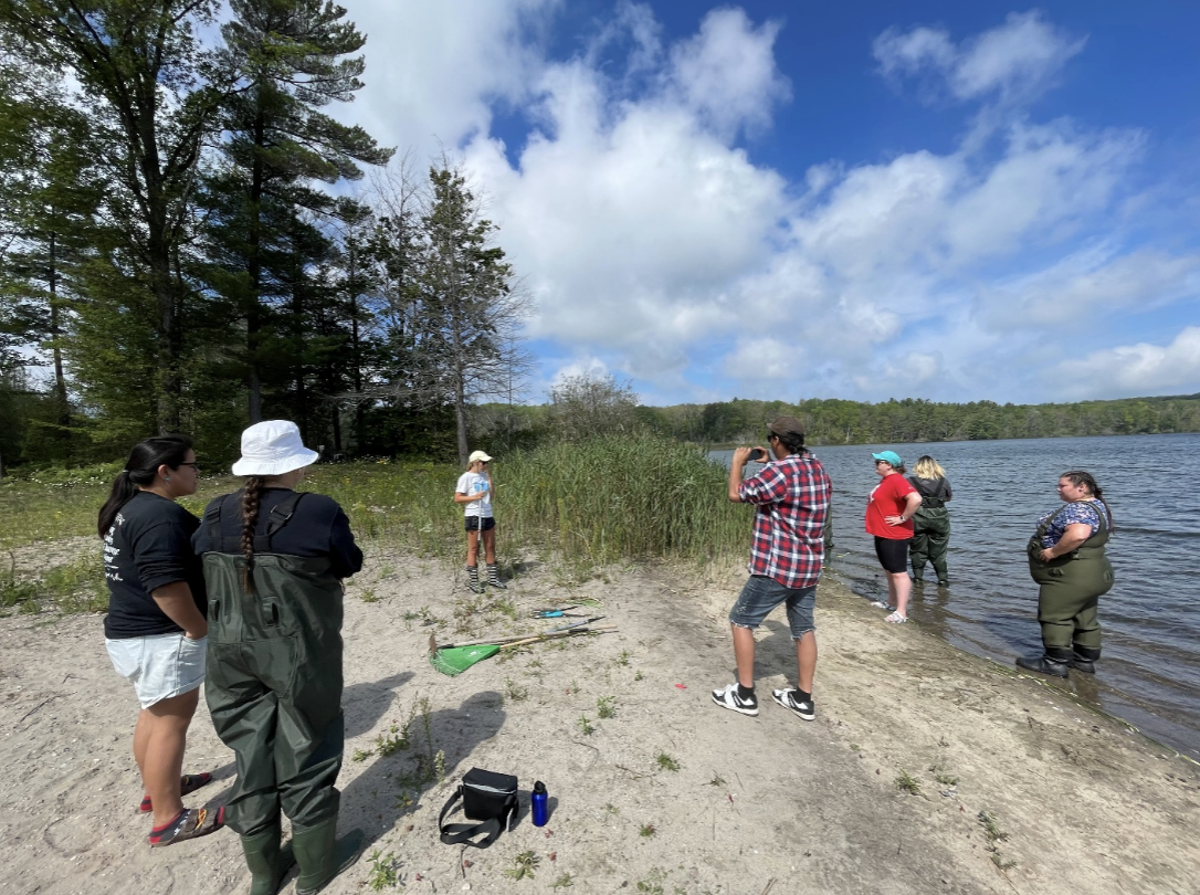 Group of people standing on a sandy lakeshore with trees and blue sky in the background, some wearing rain gear and others in casual clothing, near water's edge, possibly on a nature walk or outdoor activity.