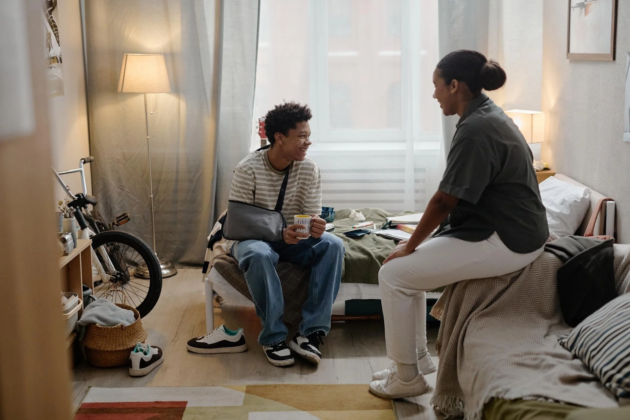 A woman talking to a man sitting on a hospital bed in a cozy room, smiling, holding a mug. There is a bicycle and a small bookshelf in the room.