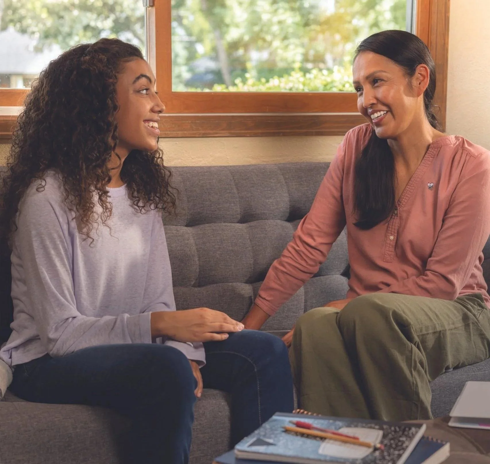 Two women, one with curly hair and one with straight hair, sitting on a couch and holding hands inside a room with a window showing trees outside, engaged in a warm conversation.