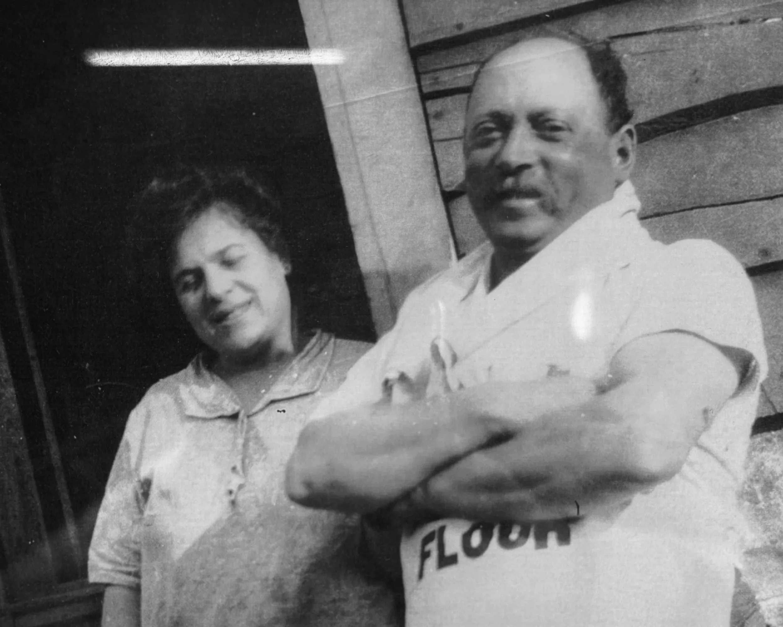 A black and white photo of a man with a mustache and a woman, both smiling, standing outdoors. The man is crossing his arms and wearing a shirt that says 'FLOUR.' The woman has short, wavy hair and is wearing a collared shirt.