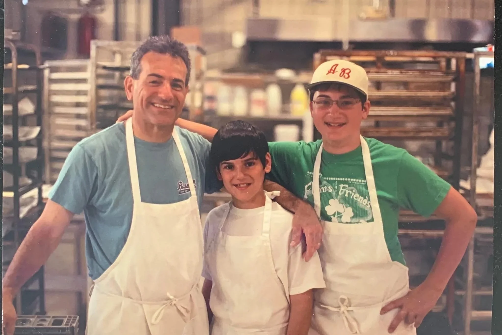 Three people standing in a kitchen or bakery, smiling and wearing aprons. Two are adult men and one is a young boy.