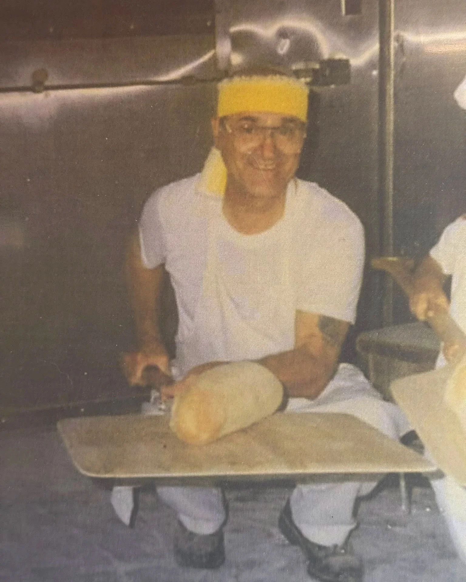 Person wearing a yellow headband and glasses, smiling while holding an Alteri's Italian loaf of bread on a wooden cutting board in a commercial kitchen.