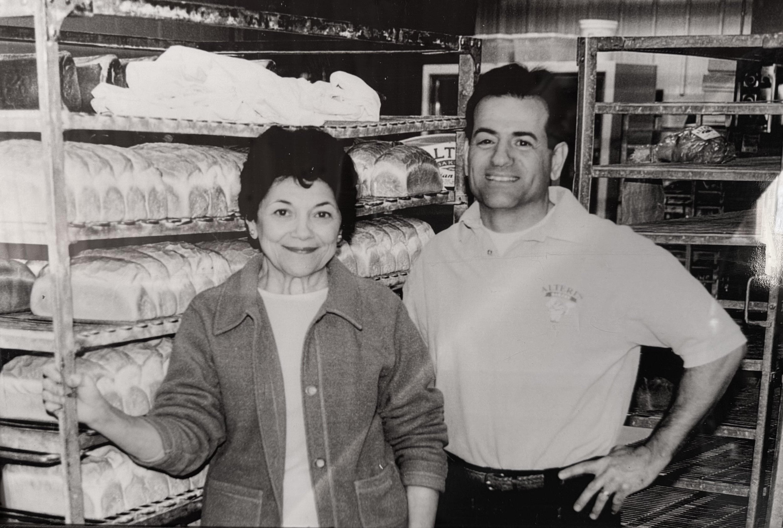 A black and white photo of a woman and a man standing in front of bread racks inside a bakery. The woman holds onto a metal rack, and both are smiling at the camera.