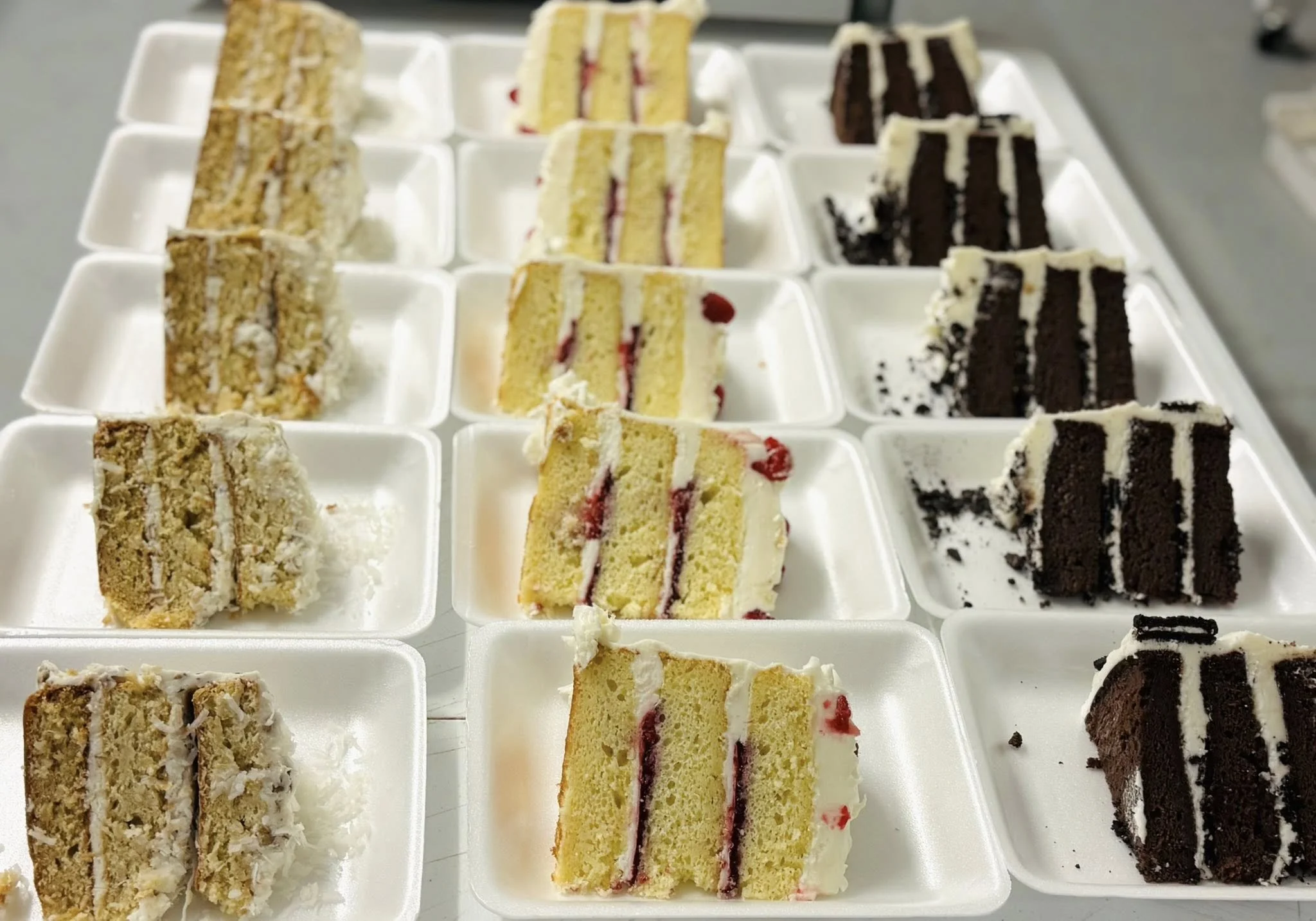 Assorted slices of cake in white foam trays featuring three types: coconut with shredded coconut on top, strawberry with pink jam filling, and chocolate with white icing and Oreo cookies.
