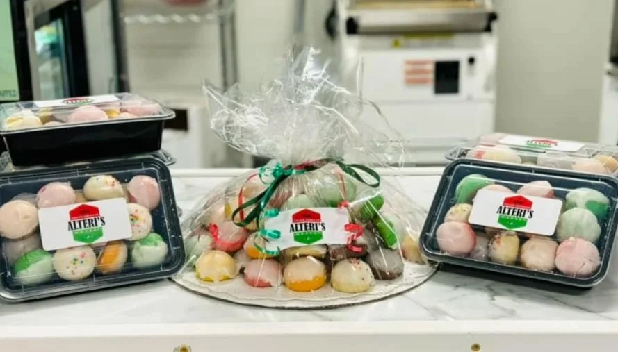 Assorted colorful macarons and treats packaged in clear plastic containers with 'Alteri's' labels, some wrapped in decorative cellophane and ribbons, on a white countertop.