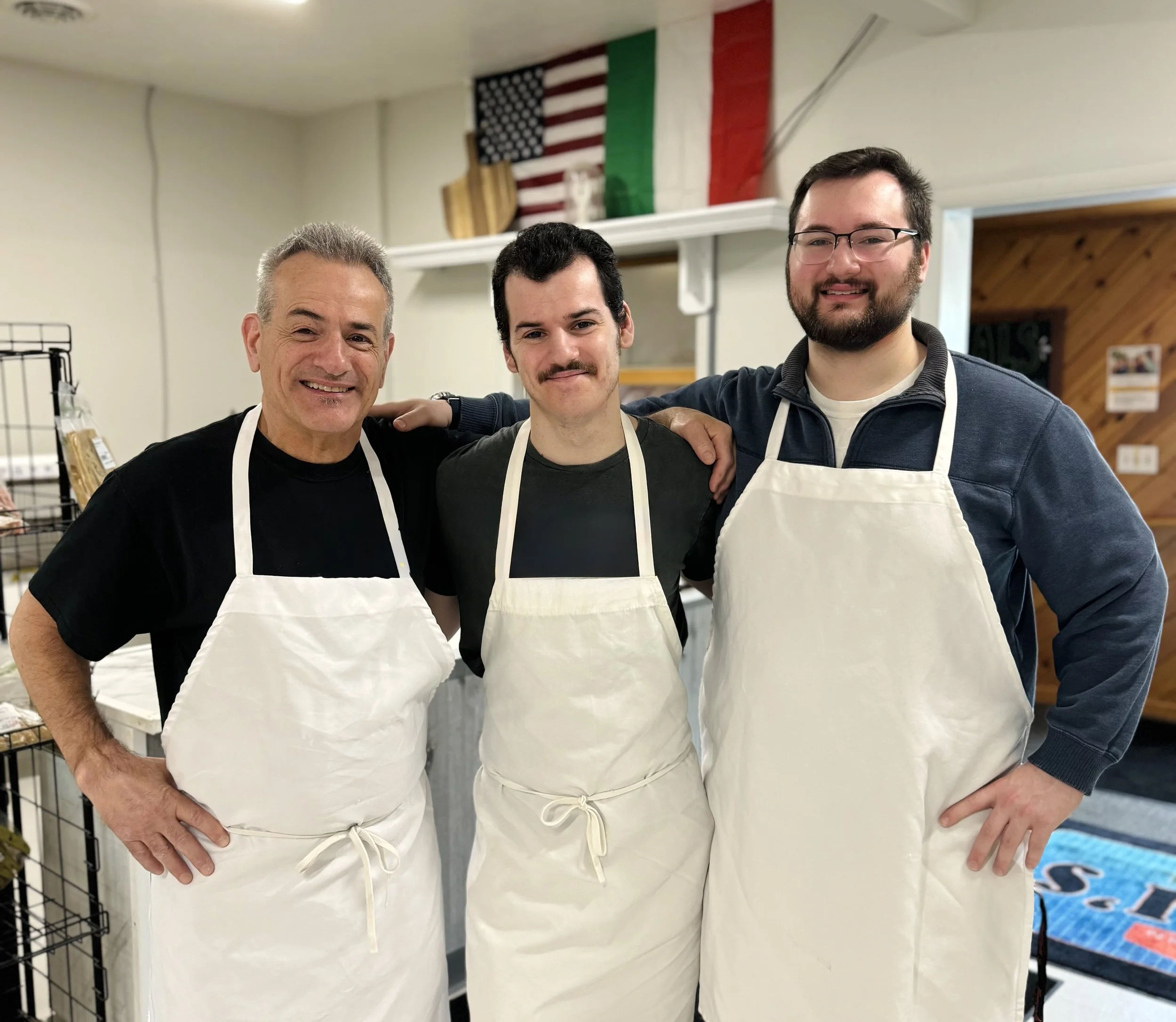 Three men wearing white aprons standing together inside a room with American, Italian, and Canadian flags in the background. They have their arms around each other's shoulders and are smiling at the camera.