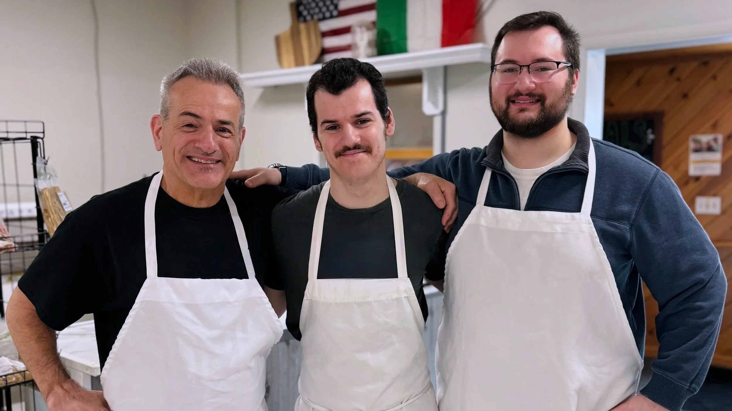 Three men standing side by side wearing white aprons inside a room, smiling at the camera. The man on the left has gray hair, the man in the middle has dark hair and a mustache, and the man on the right has dark hair and glasses. There is an American flag and Italian flag hanging behind them.