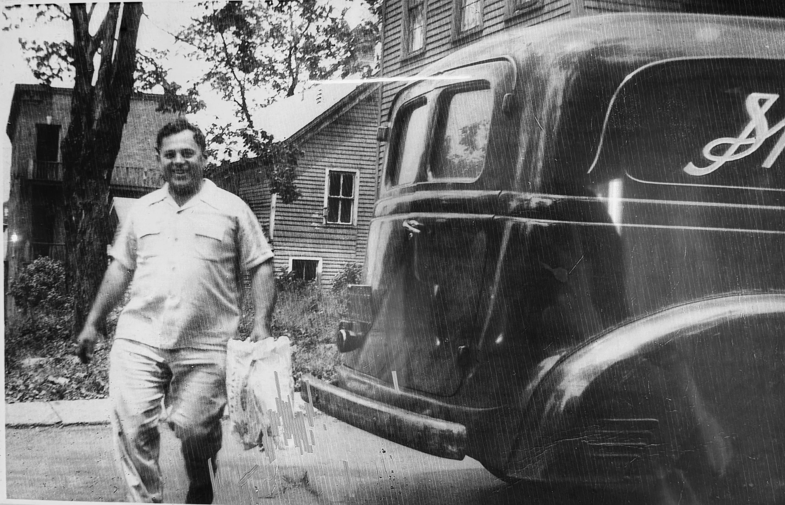 A young man walking on a sidewalk next to a black vintage truck with Spaziani's Bakery its side, in front of houses with trees in the background.