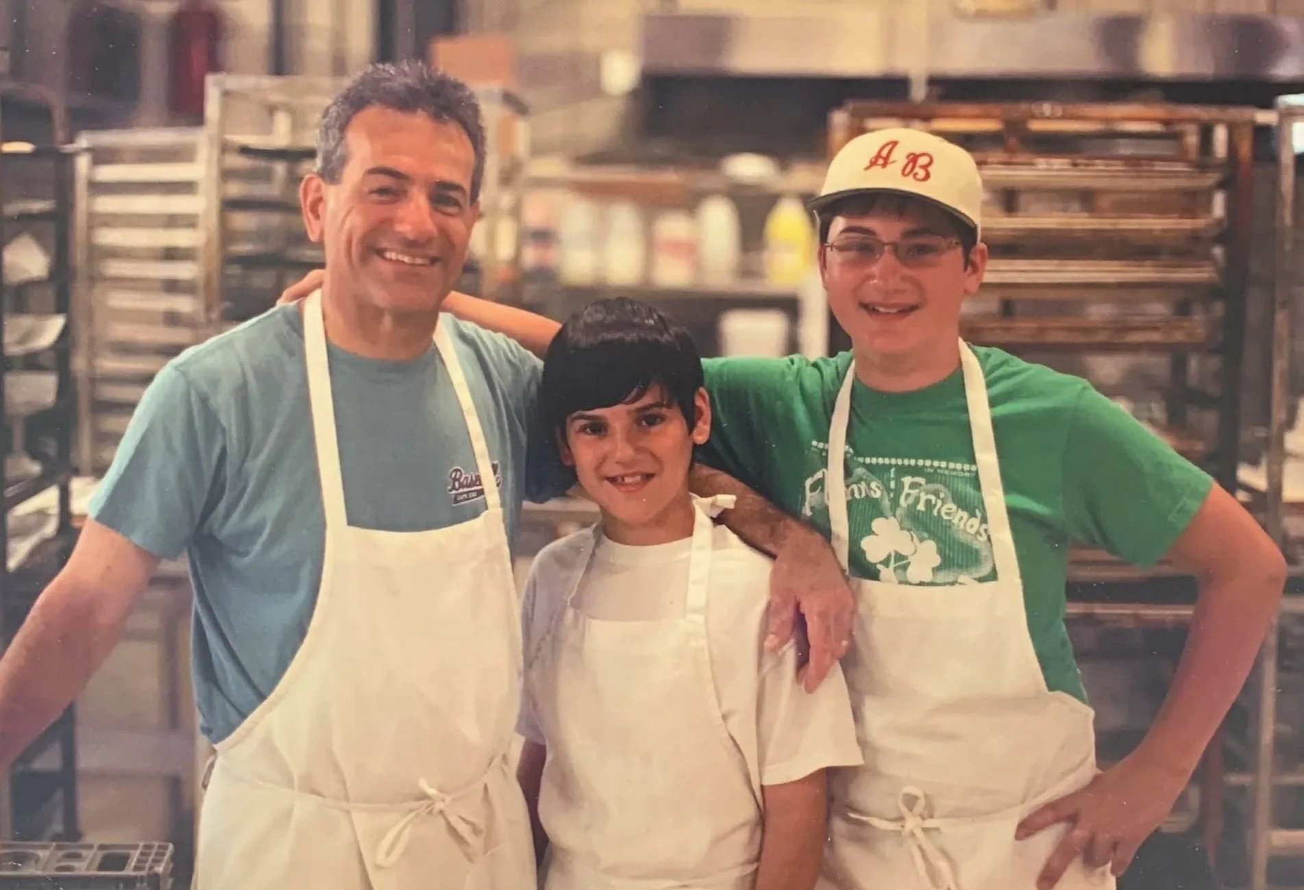 Three people, two adults and one child, in a bakery or kitchen setting, all smiling and wearing white aprons. The man on the left has short dark hair and a light blue t-shirt. The boy in the middle has dark hair and a white t-shirt. The man on the right is wearing glasses, a green t-shirt, and a baseball cap with red embroidery.