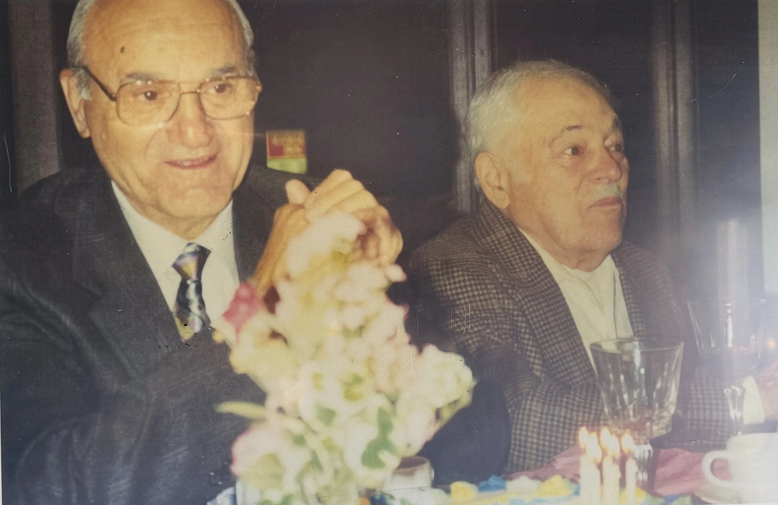 Two elderly men sitting at a table during a celebration, with one holding a lit candle on a cake and a flower arrangement in front of them.