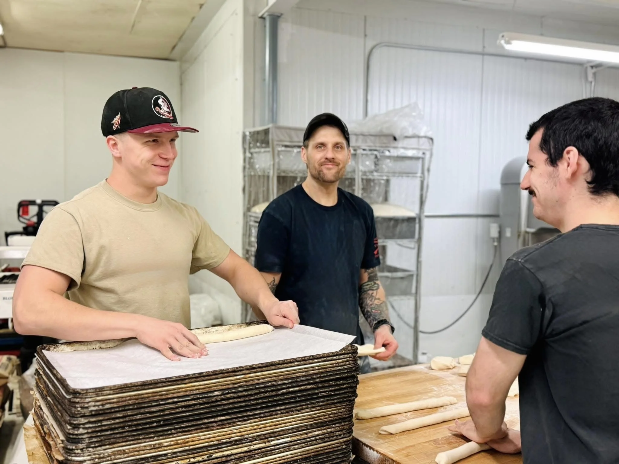 Three men in a bakery or dough preparation area, with one man in a beige shirt and black cap rolling dough, another in a black shirt with tattoos on his arms, and a third man in a black shirt smiling, with stacks of dough sheets and rolling pins on the table.