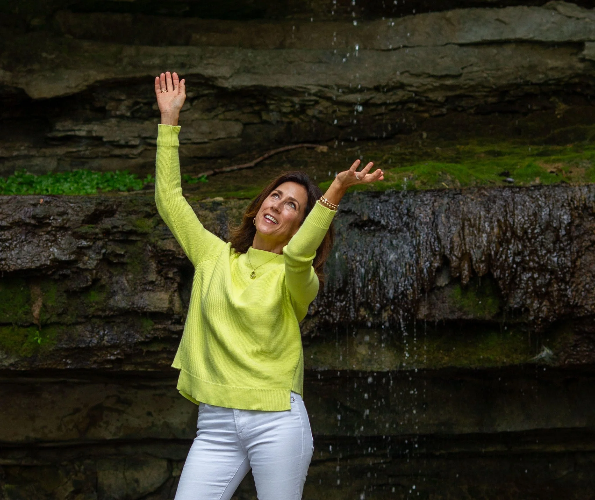 Woman in a bright green sweater and white pants standing by a rocky waterfall, raising her arms and smiling — The Boob Project empowerment portrait.