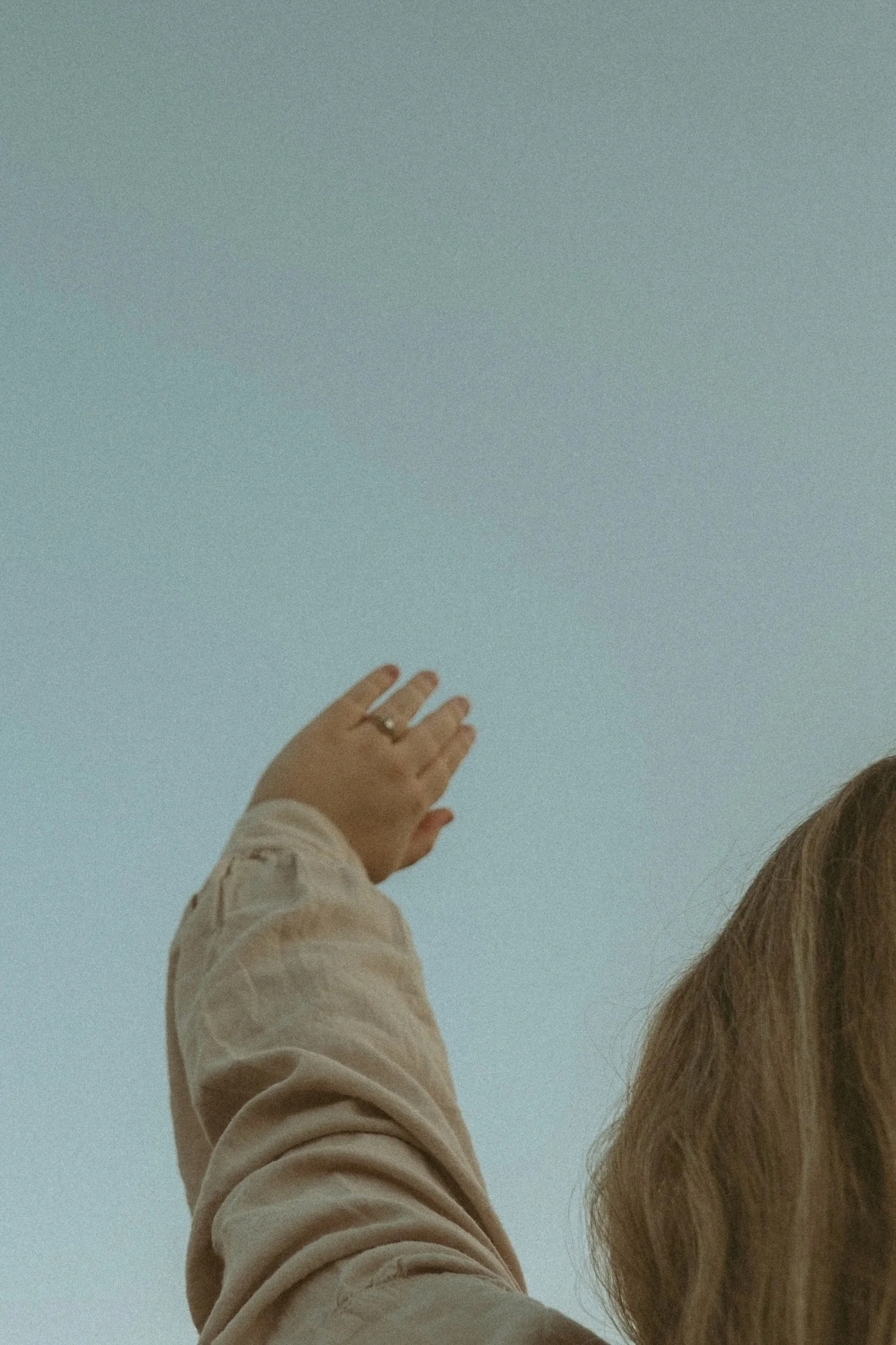 A person's hand with a wedding ring, reaching toward a pale blue sky, partially visible with long hair on the right side of the image.