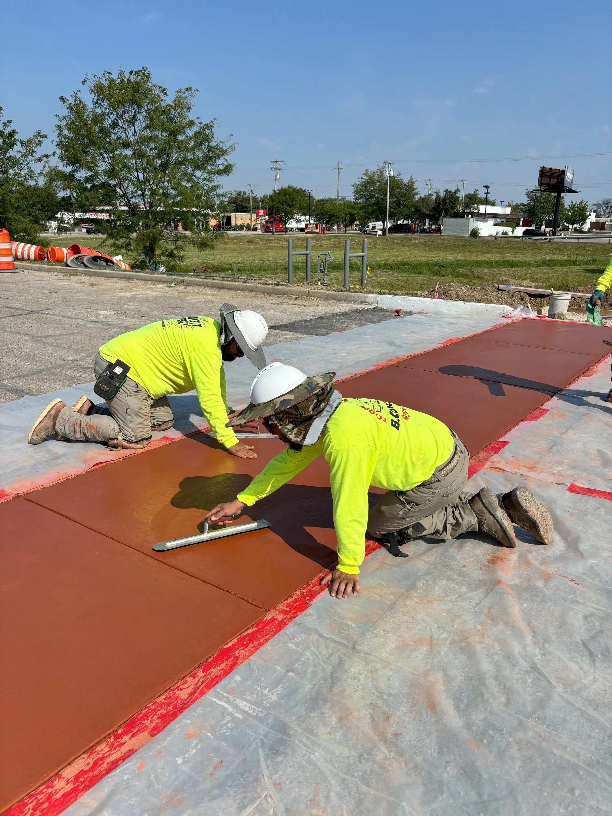 Construction workers wearing safety gear install a new sidewalk on a street, with one worker using a tool to smooth the surface.