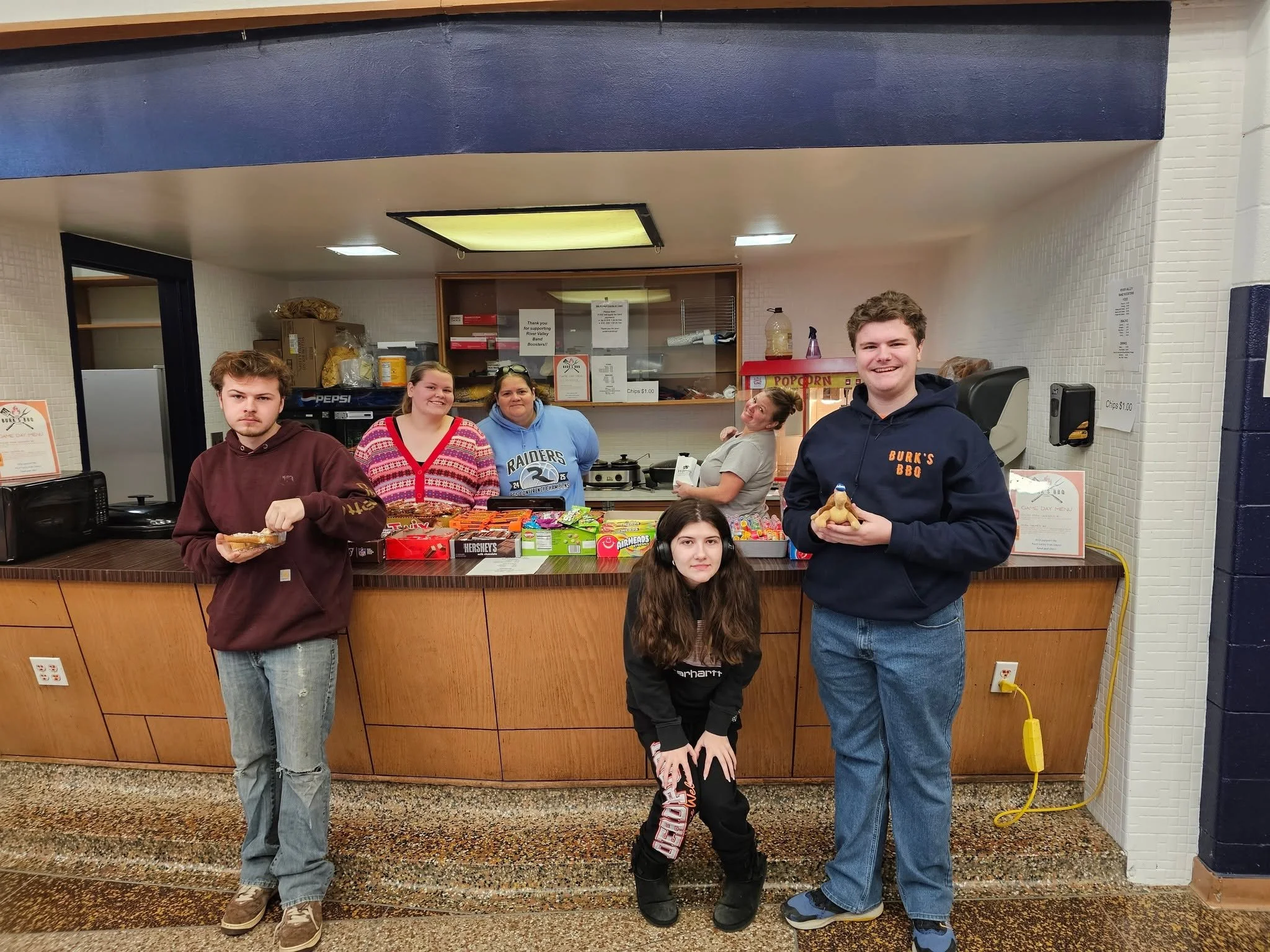 Group of five people standing behind a counter at a food stand or kiosk, with candy, snacks, and popcorn visible on the counter. They are smiling and looking at the camera, and the background includes a food service area with signs and supplies.