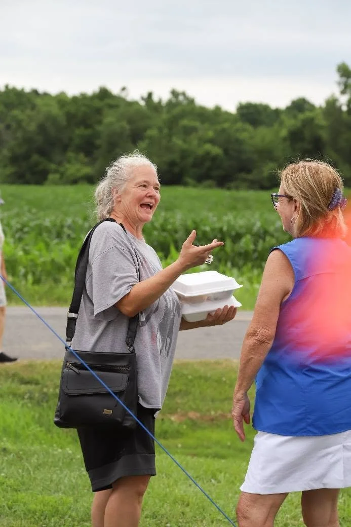 Two women having a conversation outdoors on a grassy area with a green field and trees in the background. One woman is holding a takeout food container and smiling, wearing a gray top and black skirt, with a black crossbody bag. The other woman is we