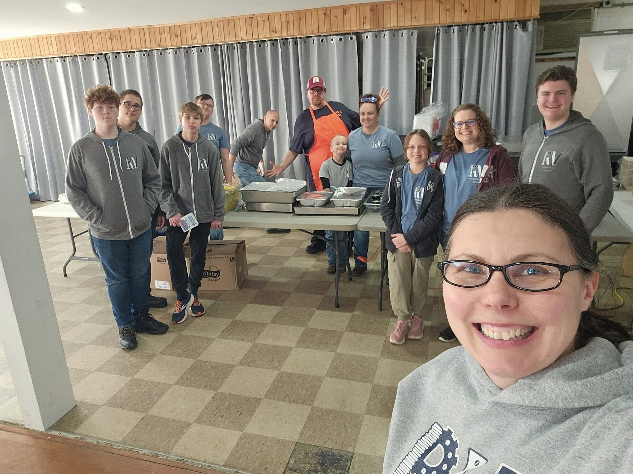 A group of children and adults standing behind a table with food trays, posing for a photo at an indoor event, with a woman in the front taking a selfie.