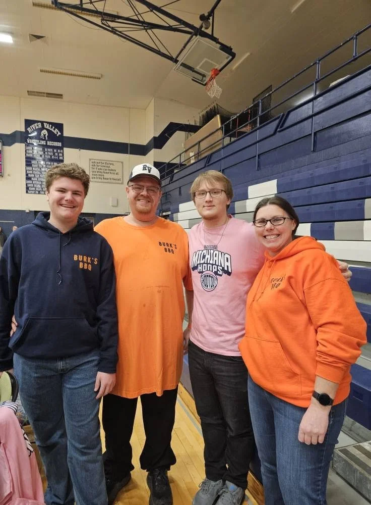 Four people standing in a gymnasium, smiling, with a blue and white bleacher in the background, and a basketball hoop hanging from the ceiling. Two are wearing Michigan Wolverines shirts, and one has a hoodie and shirt with 'Bucky's BBQ'.