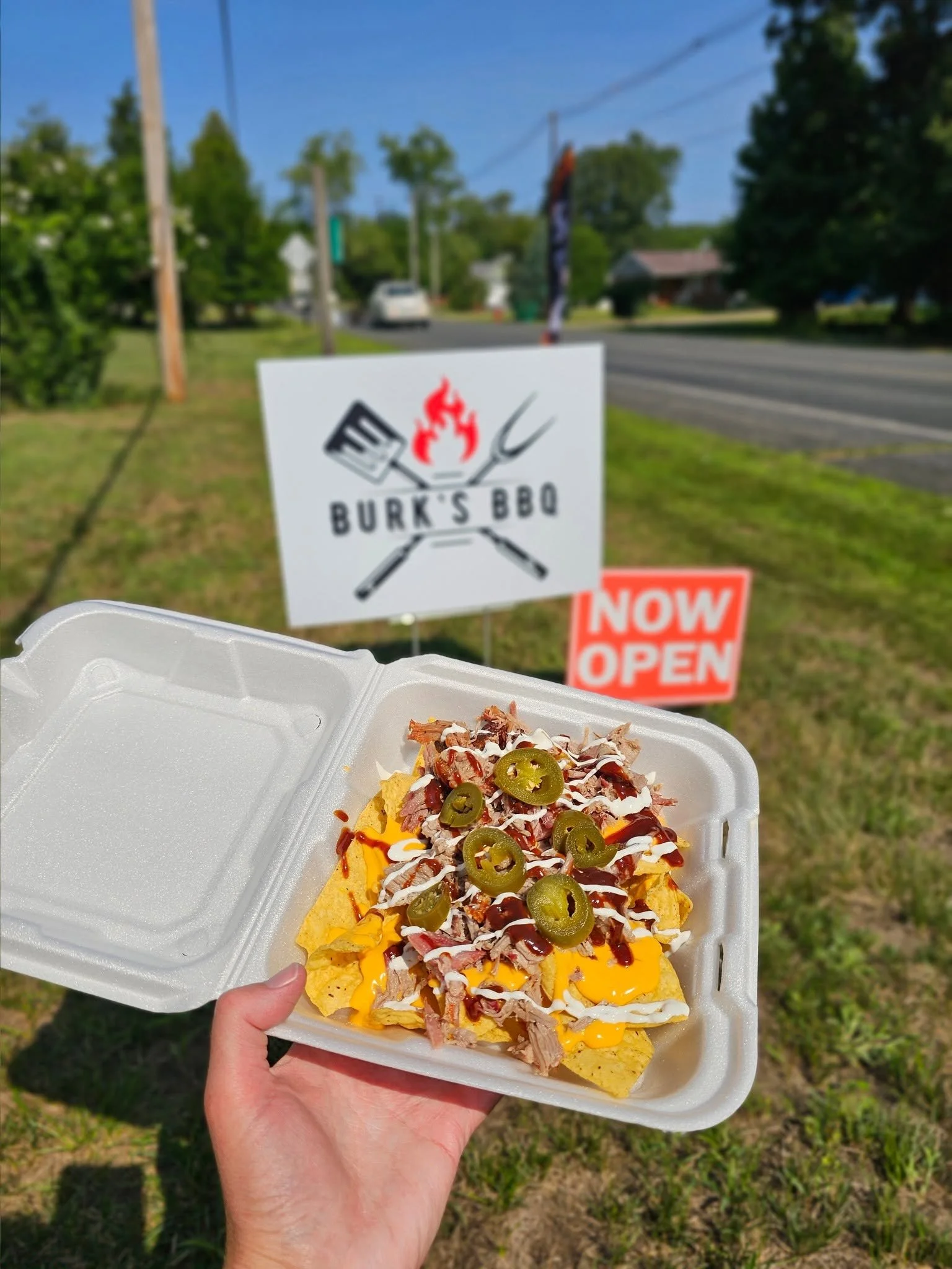 A hand holding a styrofoam food container with nachos topped with jalapenos, cheese, sour cream, pulled pork meat, and sauce, in front of a blurred outdoor scene with trees, street, and signs advertising a BBQ restaurant that is now open.