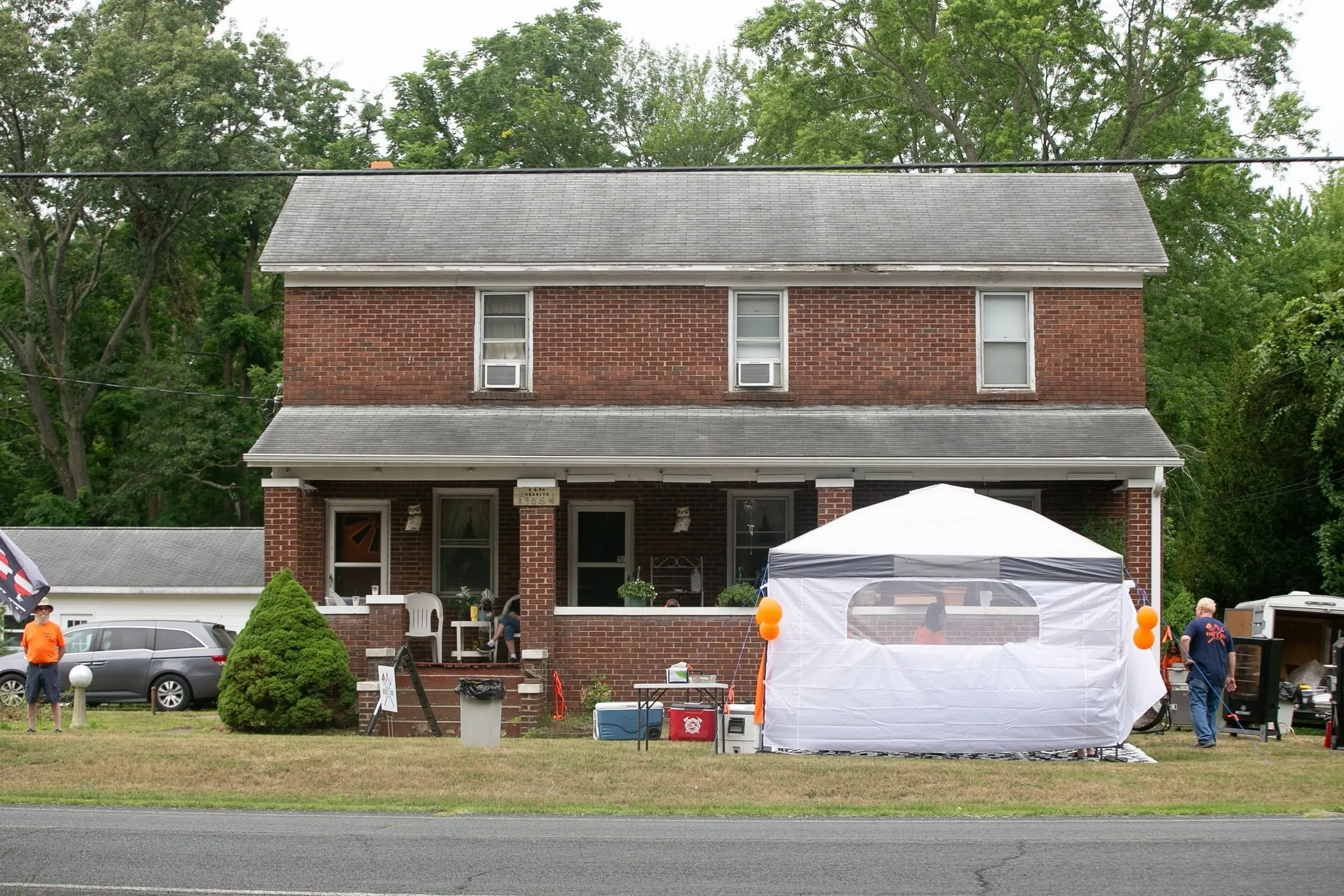 A two-story brick house with trees in the background and a driveway fronting the house. There are tents and balloons in the yard, indicating a gathering or event, with people standing around.