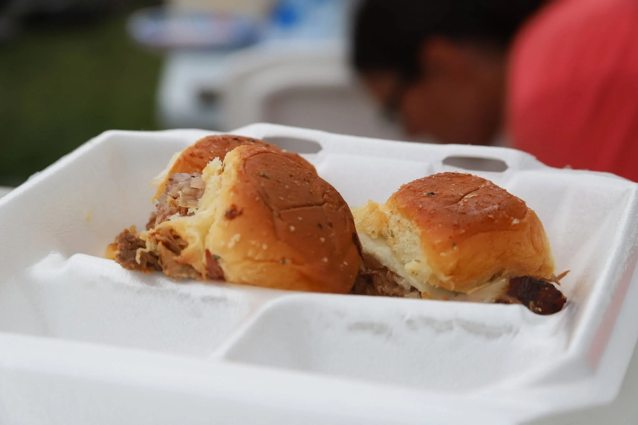 Close-up of two pork sliders food in a white foam takeout container, with one partially broken open showing the shredded meat filling inside.