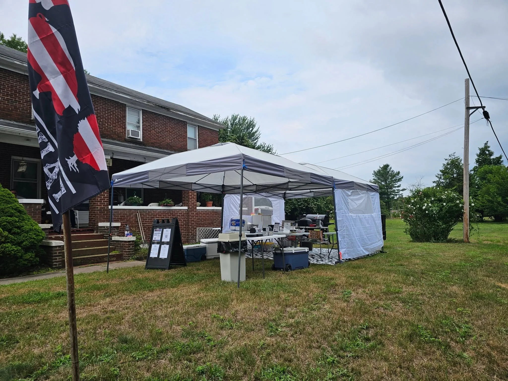 An outdoor setup in front of a brick house with a white canopy tent and various tables and items underneath. A black flag with red, white, and black elements is partially visible on the left side. The area is grassy with some trees and shrubs, and a 