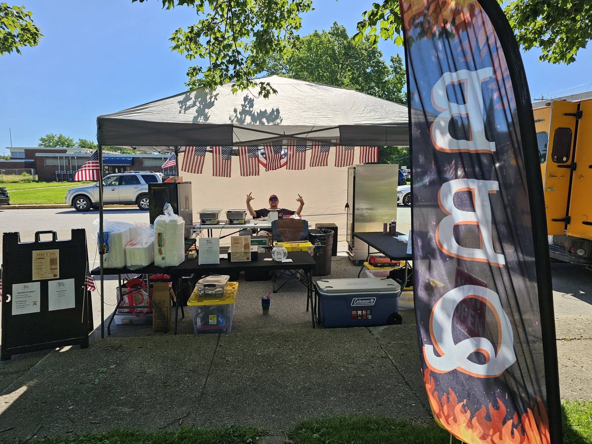 A food stand under a canopy decorated with American flags, with a man sitting behind the counter , and a large banner on the right side of the image.