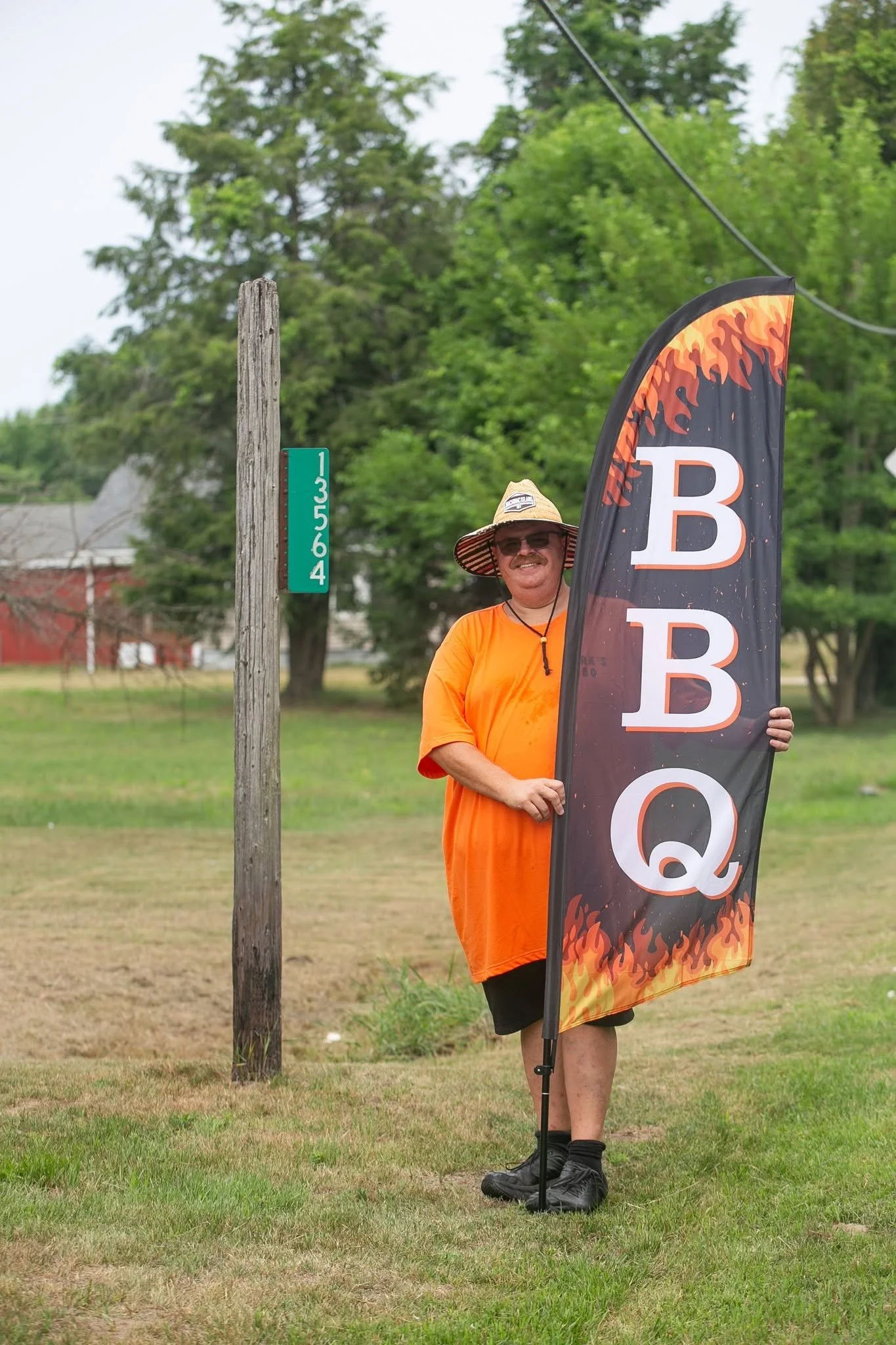 A smiling man wearing sunglasses, a wide-brimmed straw hat, an orange T-shirt, and black shorts standing outside on grass, holding a QA flag with flames and a large letter B on it, near a wooden pole with a green numbered marker.