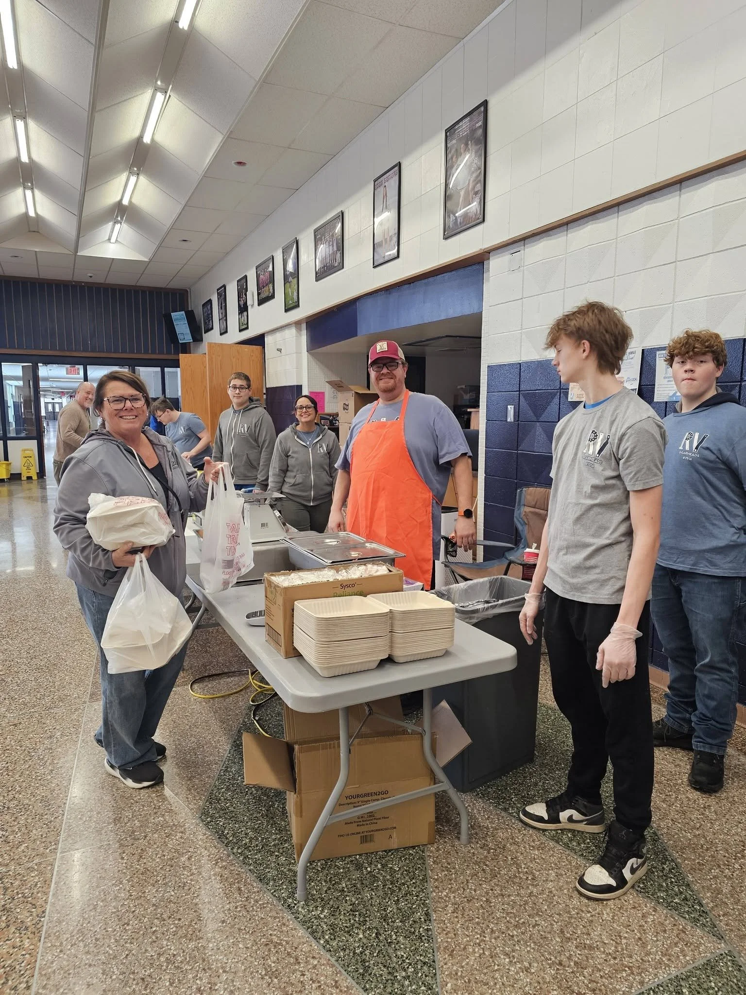 People standing around a temporary food service table in a public indoor space, some wearing gloves. There are boxes and food items on the table, with a woman smiling at the camera and others working or waiting in line. Burk's BBQ fundraiser.