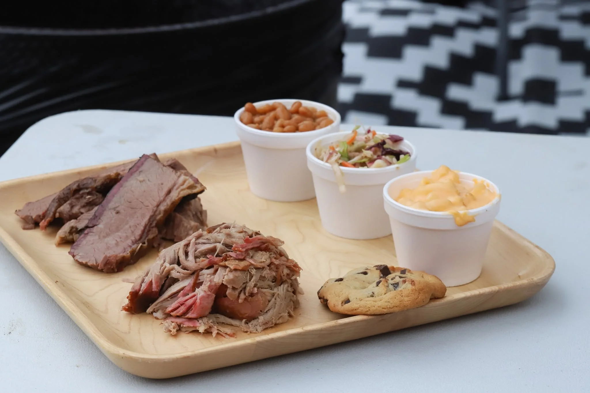 A wooden tray with sliced brisket, pulled pork, and several small cups of sides including baked beans, coleslaw,  and a chocolate chip cookie.