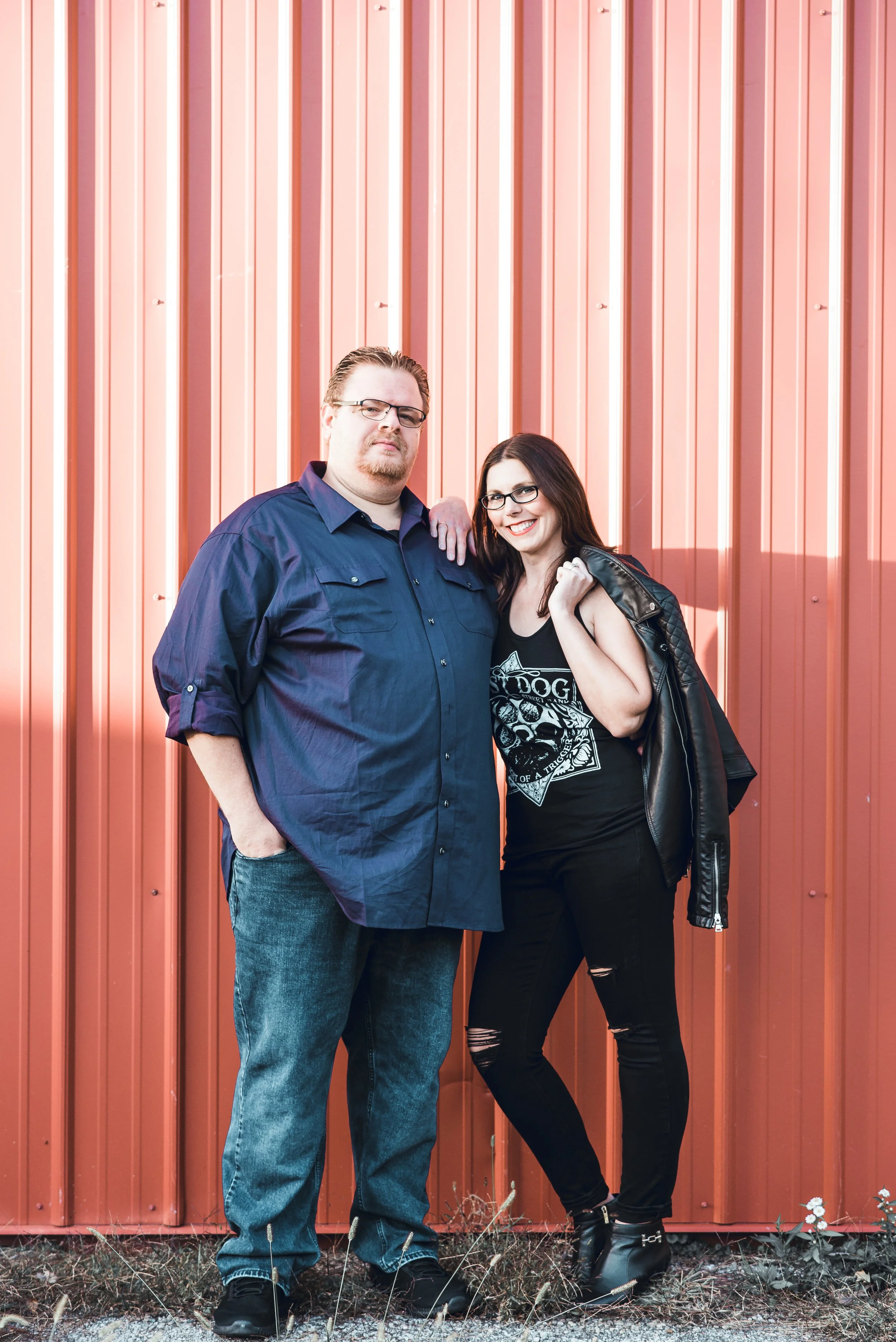 A man and woman stand together outside in front of a red corrugated metal wall. The man is wearing glasses, a dark blue button-up shirt, and jeans. The woman has long dark hair, glasses, and is dressed in a black T-shirt with a graphic design, ripped black jeans, and a black leather jacket.