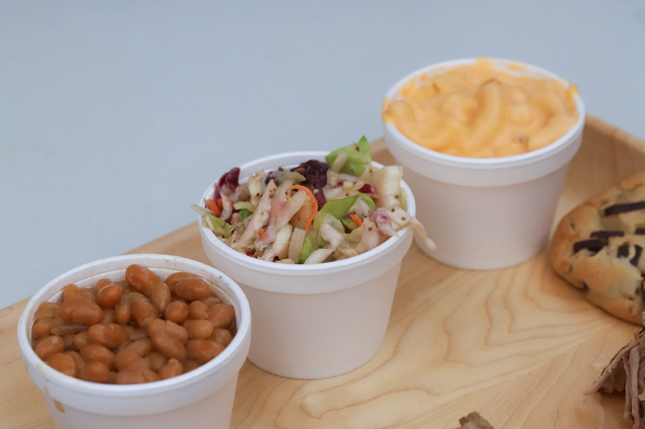 Three small white cups filled with different food items on a wooden surface, including baked beans, coleslaw, macaroni and cheese, and a cookie.