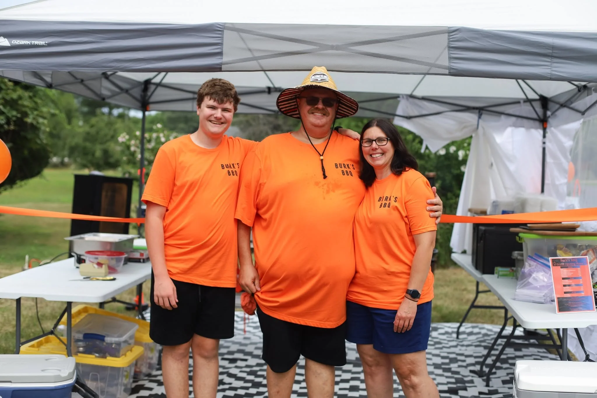 Three people wearing matching orange T-shirts are standing under a canopy at an outdoor event. They are smiling and appear happy, with the man and one woman having their arms around each other. 
