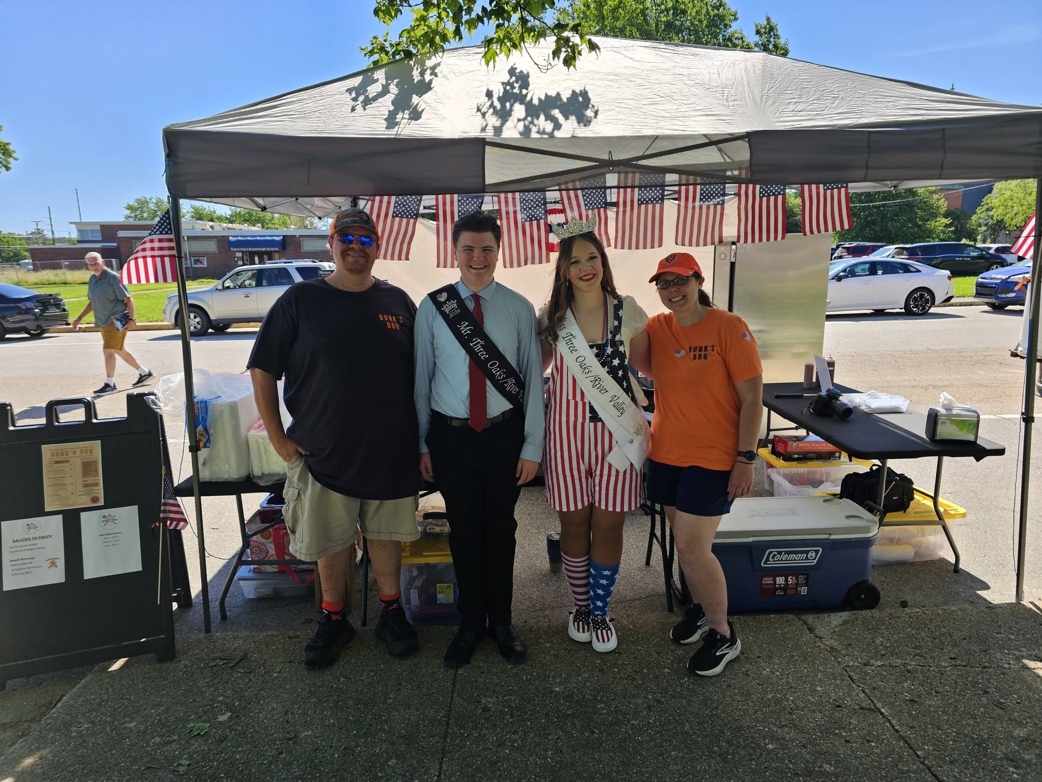 Four people standing under a small outdoor tent decorated with American flags, at a celebration event. Burk's BBQ Flag Day Three Oaks, MI